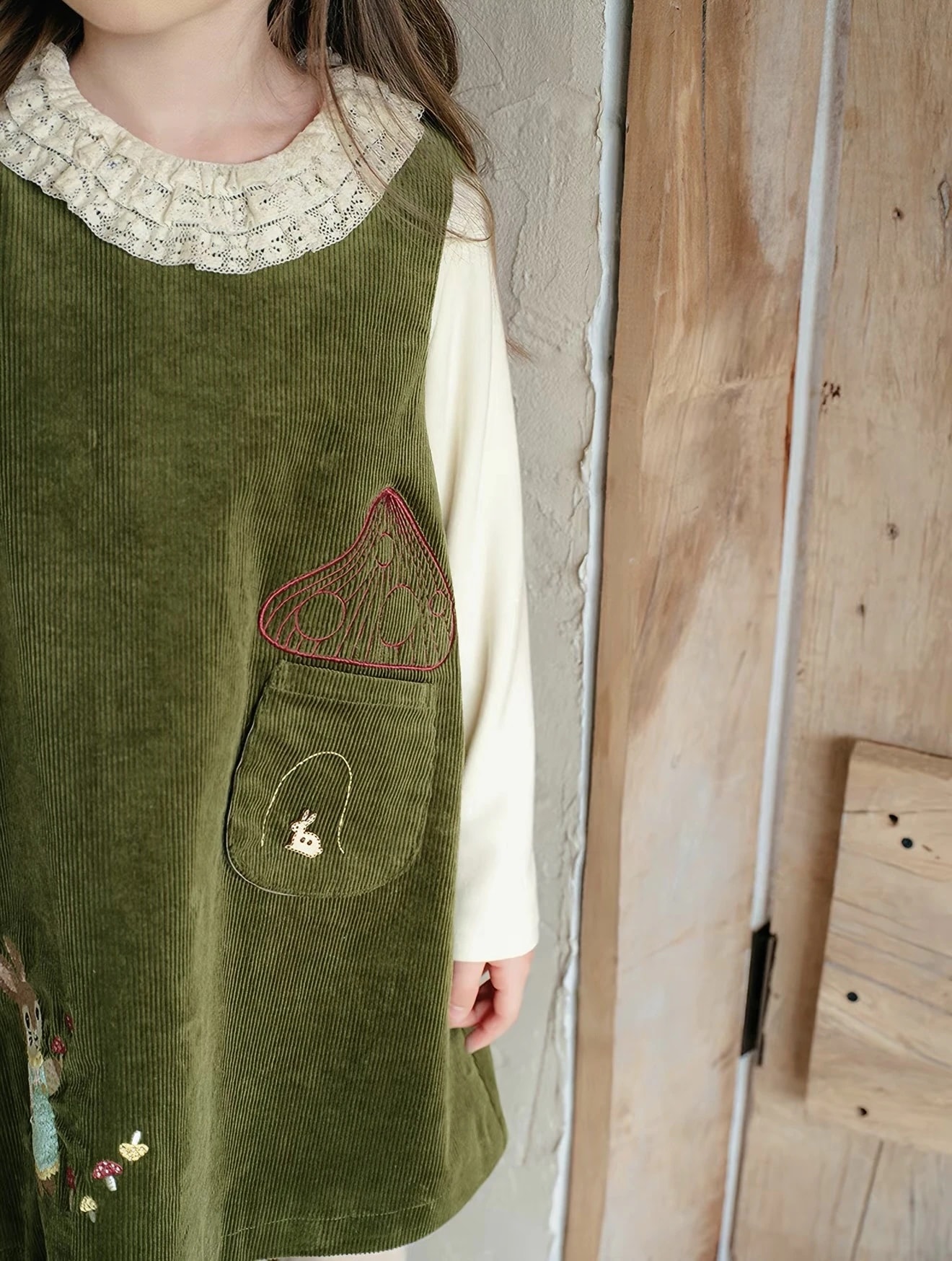 Detail Madeleine Dress: saku korduroi dengan jahitan jamur merah dan kelinci kecil, serta kerah renda blus dalam.