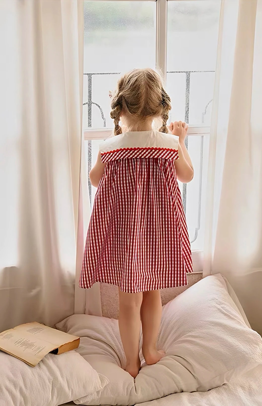 Tampak belakang anak perempuan memakai Rosalie Dress di dekat jendela, terlihat kerah lebar dengan list gingham dan pita gelombang merah.