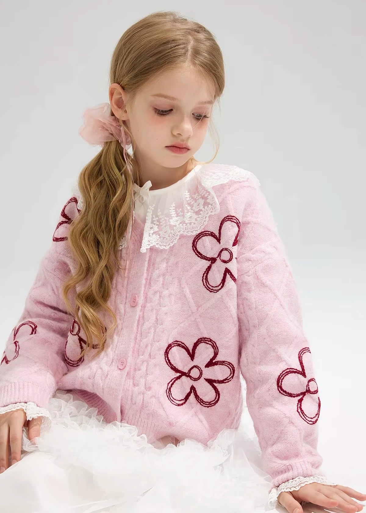 Duduk memakai Clemence Cardigan pink di atas blus berkerah renda, terlihat bordir bunga burgundy di bagian depan dan lengan.