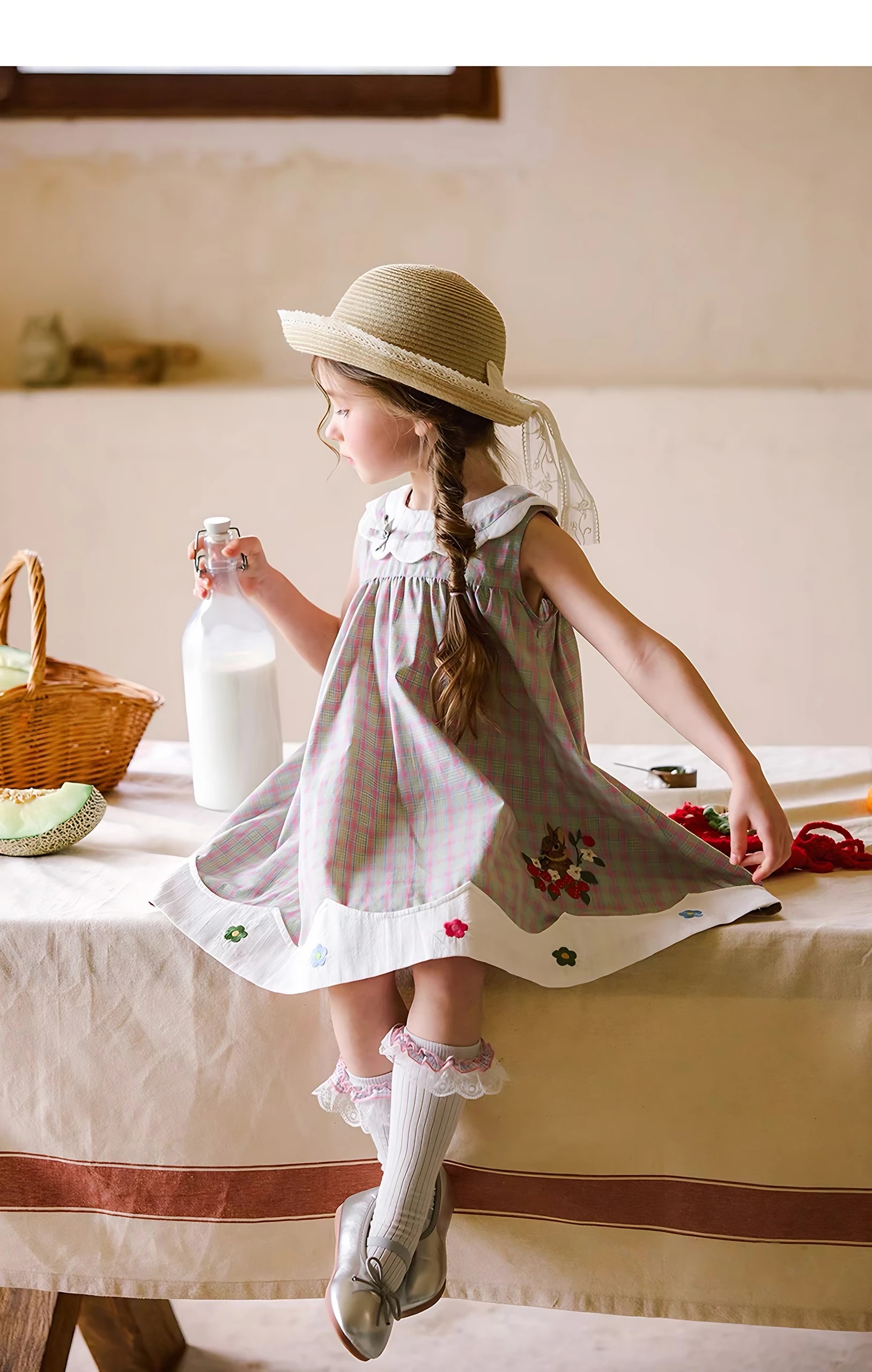 Tampak samping anak perempuan duduk di meja memakai Celestine Dress, terlihat rok kotak-kotak mengembang dan bagian bawah putih bergelombang.