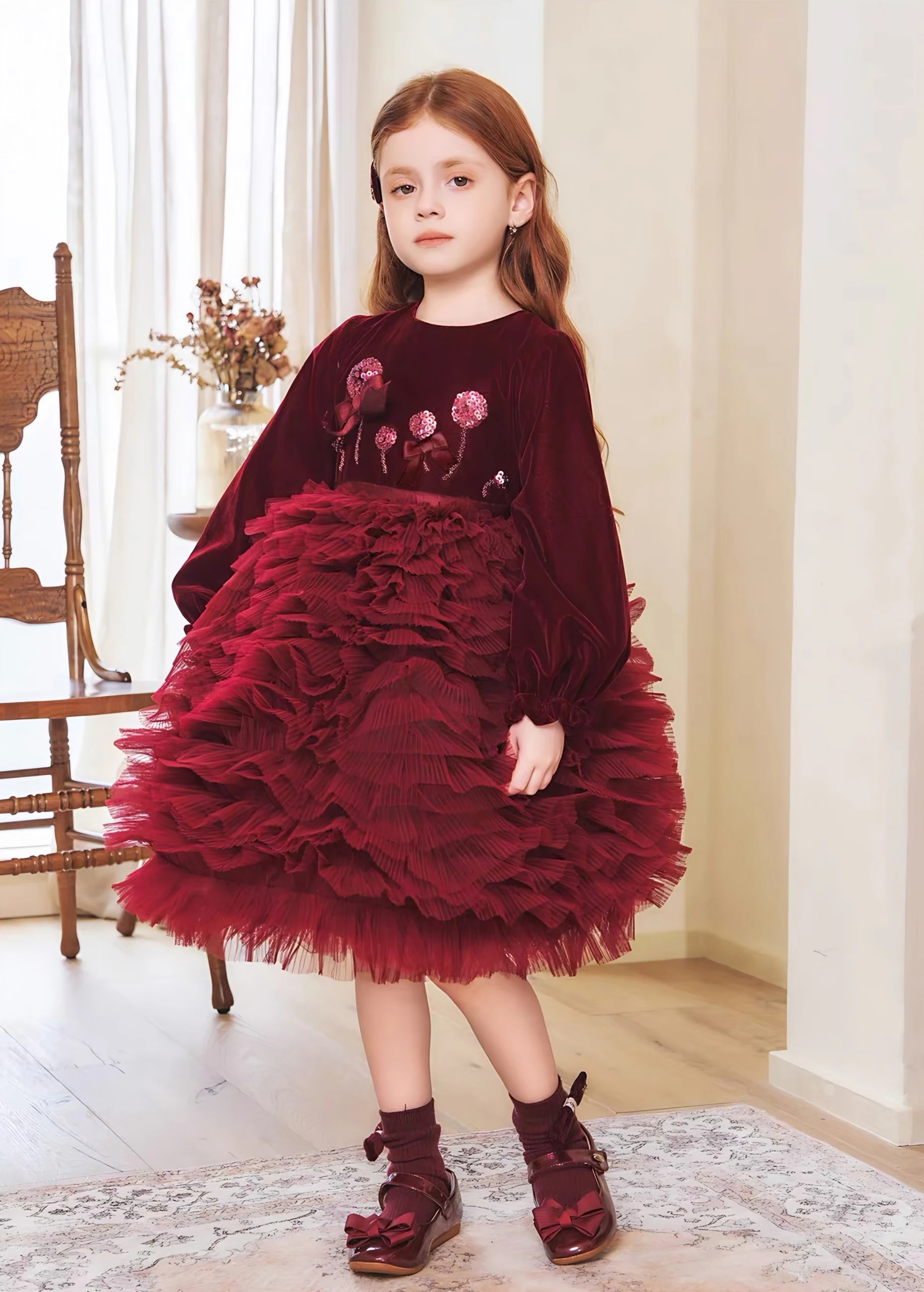 Anak perempuan berdiri memakai Eleonore Dress burgundy, terlihat lengan panjang mengembang dan bunga payet berpita kecil di atasan.