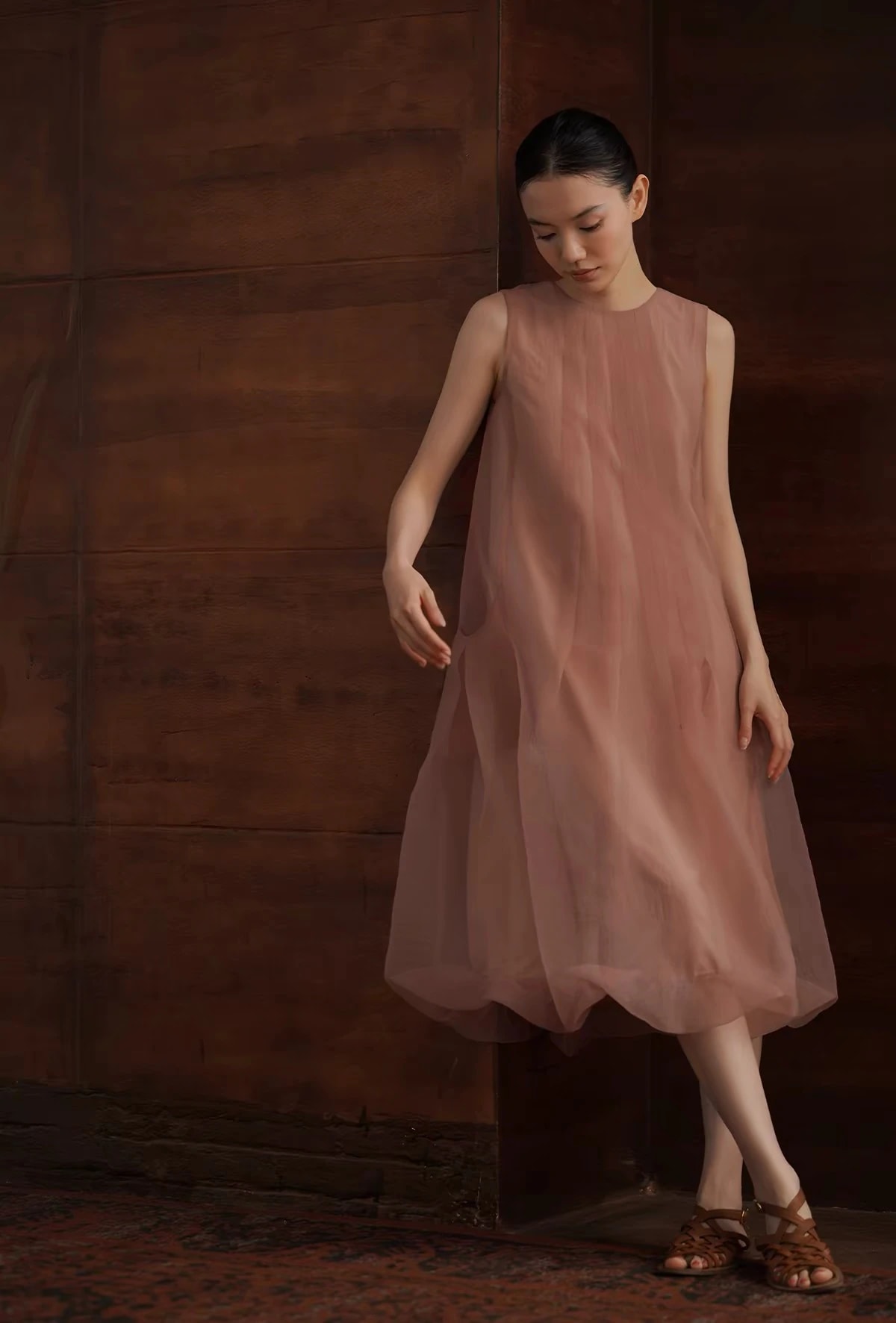 A woman in the dusty pink Solene Dress, a Christmas outfit, leaning on a dark wood wall as the sheer gauze falls to a rounded hem.