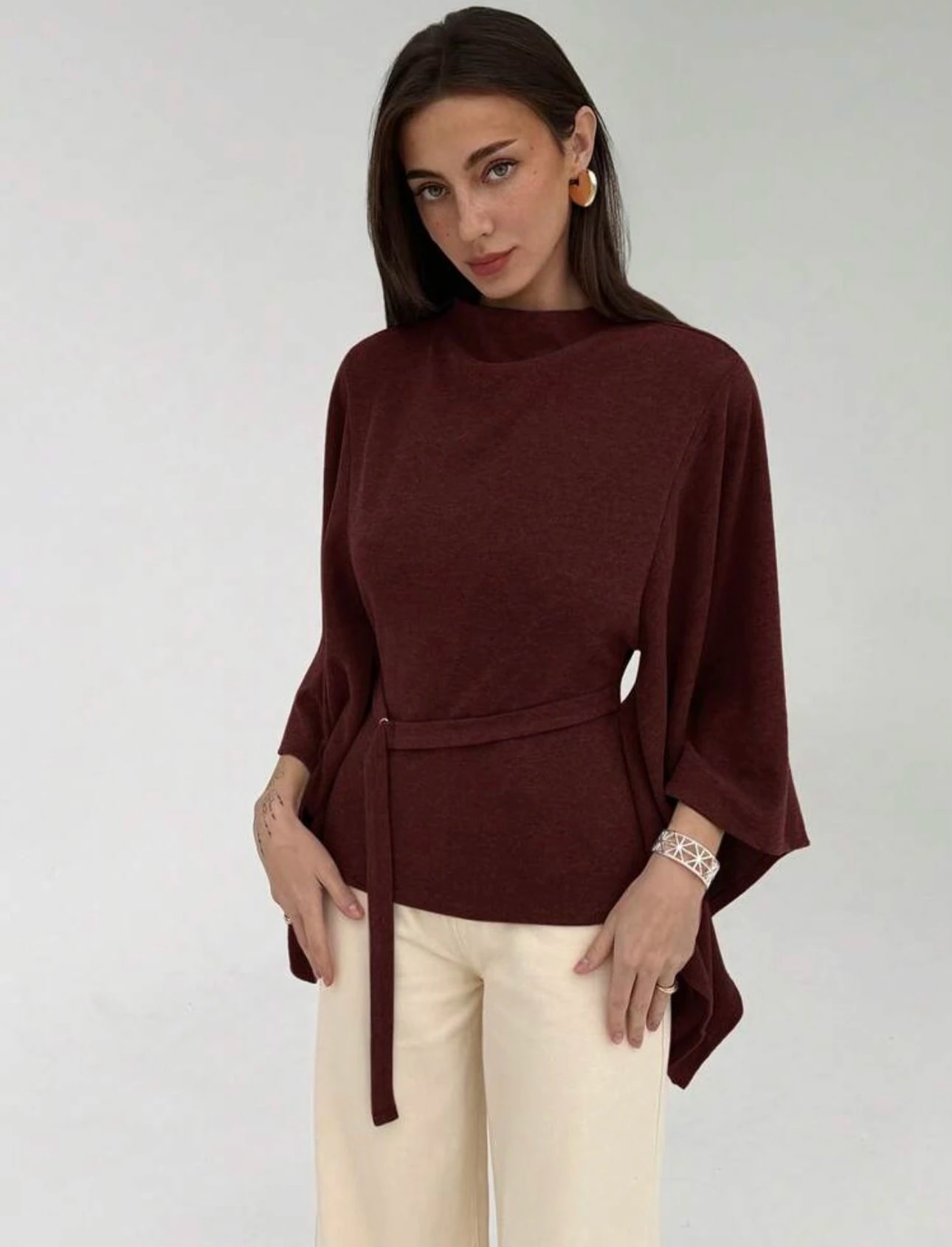 A woman in the maroon Liliane Top, a Christmas outfit, with a stand collar, wide batwing sleeves and a tie at the waist.