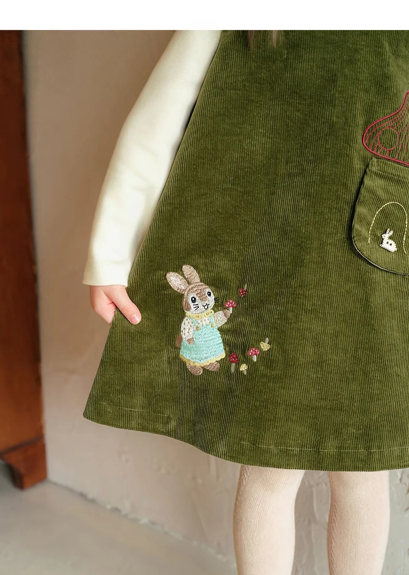 Detail bordir kelinci di dekat bagian bawah Madeleine Dress korduroi hijau zaitun, dengan jamur-jamur kecil di sampingnya.