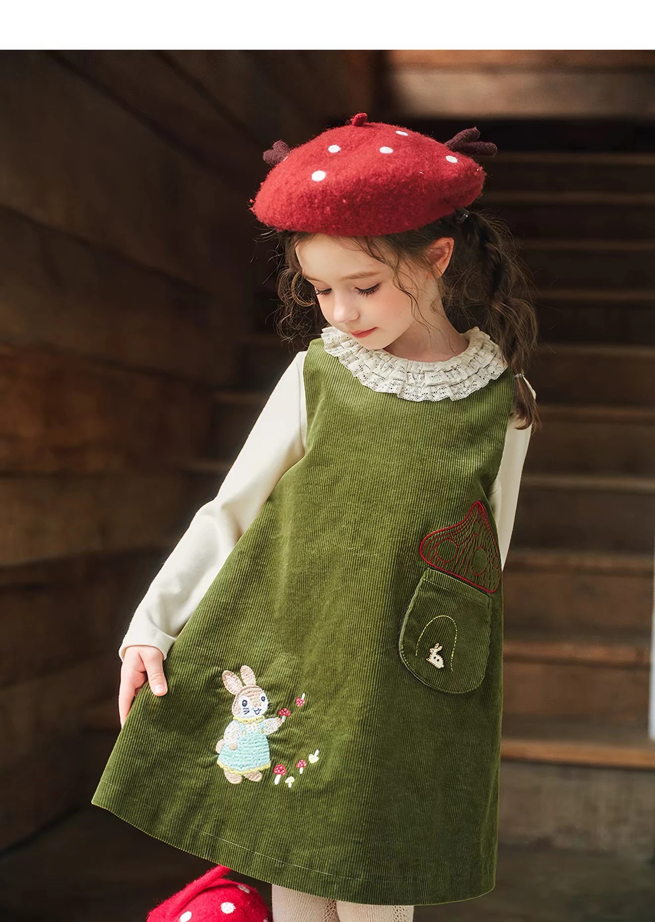 Anak perempuan menunduk melihat Madeleine Dress yang dipakainya, terlihat pinafore model A-line dan kerah renda rimpel blus dalam.