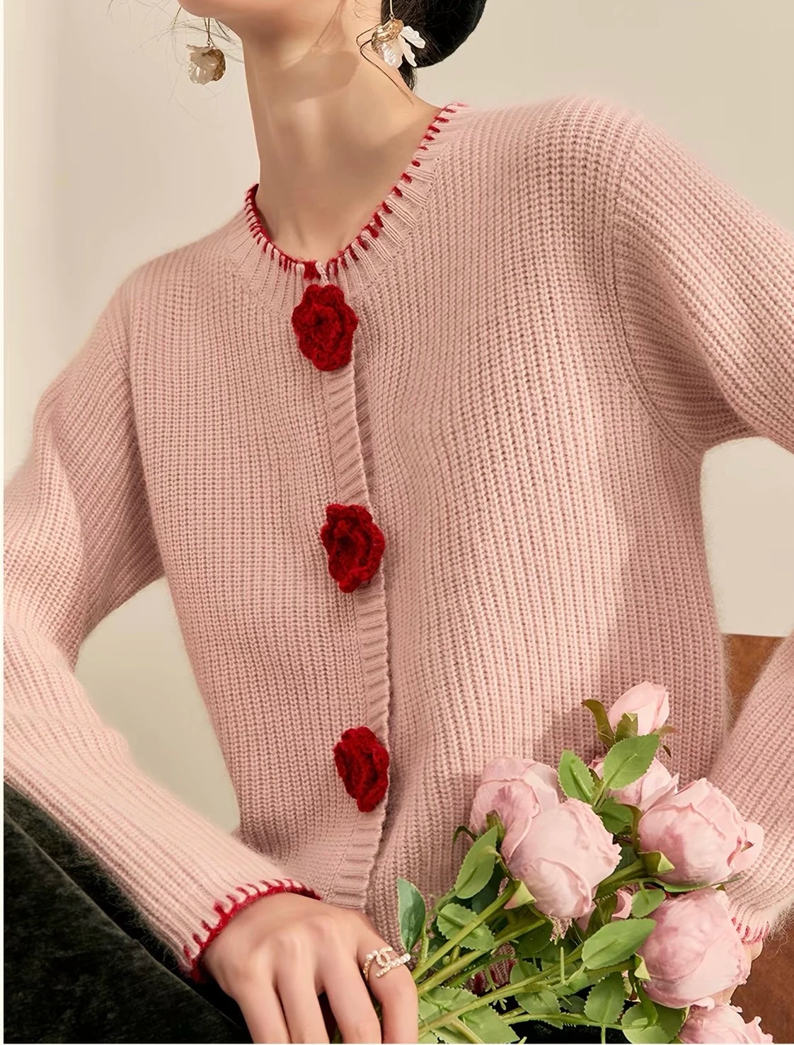 Detail depan Rosette Cardigan: tiga kancing mawar rajut merah dan kerah bulat berjahitan tepi, di samping mawar pink.